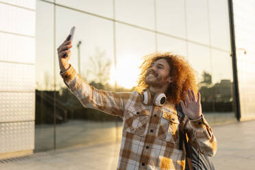 Happy man waving on video call through smart phone near building - JCCMF11148