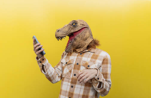 Man wearing dinosaur mask and using smart phone in front of yellow wall - JCCMF11164