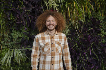 Happy redhead man standing in front of plants - JCCMF11170