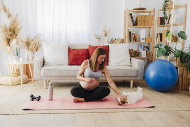 Smiling pregnant woman sitting cross-legged petting dog on exercise mat at home - EBBF08528