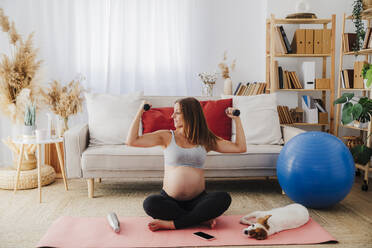 Schwangere Frau, die mit Hanteln im Schneidersitz auf einer Yogamatte zu Hause trainiert - EBBF08530