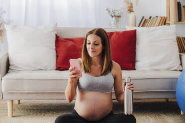 Schwangere Frau mit Wasserflasche und Smartphone im heimischen Wohnzimmer - EBBF08532