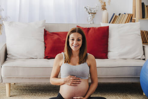 Lächelnde schwangere Frau sitzt mit Händen auf dem Bauch im Wohnzimmer zu Hause - EBBF08533
