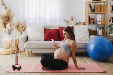 Pregnant woman using smart phone on exercise mat in living room at home - EBBF08536
