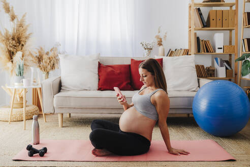 Schwangere Frau benutzt Smartphone auf Übungsmatte im Wohnzimmer zu Hause - EBBF08536