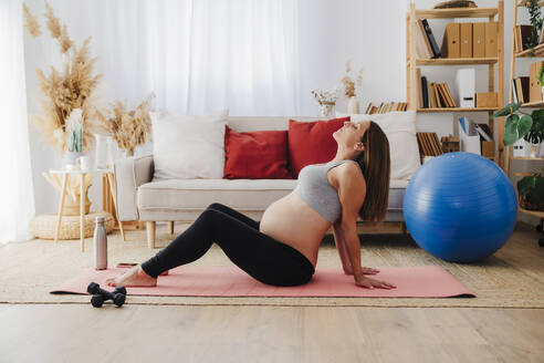 Schwangere Frau, die zu Hause auf einer Übungsmatte liegend Yoga praktiziert - EBBF08540