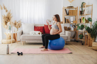 Pregnant woman using smart phone sitting on fitness ball in living room at home - EBBF08541