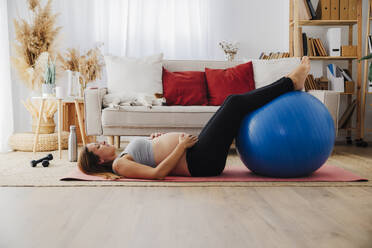 Schwangere Frau liegt auf Yogamatte mit Fitnessball zu Hause - EBBF08546