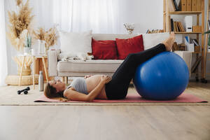 Pregnant woman lying on yoga mat with fitness ball at home - EBBF08546