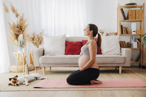 Pregnant woman kneeling with hands on stomach meditating near dog in living room - EBBF08550