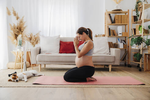 Schwangere Frau kniend auf Matte meditierend in der Nähe des Hundes im Wohnzimmer - EBBF08551