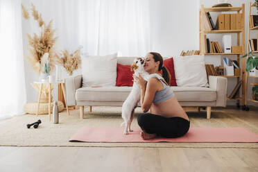 Schwangere Frau spielt mit Hund auf Übungsmatte sitzend zu Hause - EBBF08555