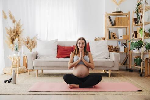 Lächelnde schwangere Frau übt Yoga auf einer Übungsmatte zu Hause - EBBF08563