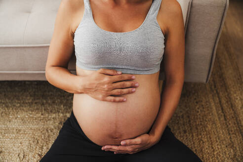 Schwangere Frau sitzt mit Händen auf dem Bauch zu Hause - EBBF08567