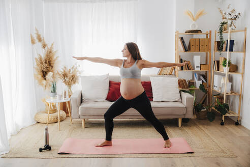 Schwangere Frau übt Krieger Pose Yoga auf Matte im Wohnzimmer zu Hause - EBBF08575