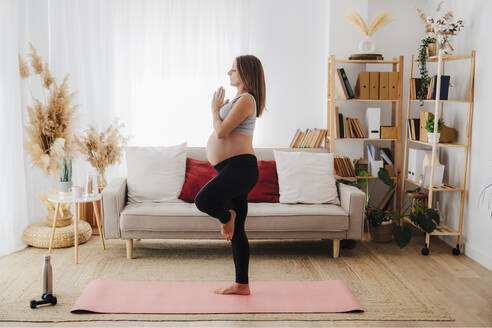 Schwangere Frau übt Baumpose Yoga auf Matte im Wohnzimmer zu Hause - EBBF08576