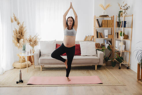 Schwangere Frau übt Baumpose Yoga auf Matte im Wohnzimmer - EBBF08577