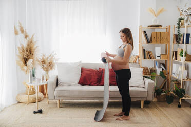 Schwangere Frau rollt Yogamatte im Wohnzimmer zu Hause - EBBF08578