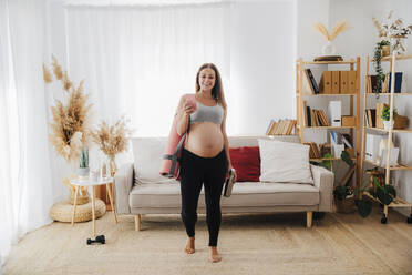 Lächelnde schwangere Frau geht mit Smartphone im Wohnzimmer zu Hause - EBBF08581