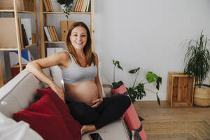 Glückliche schwangere Frau sitzt zu Hause auf dem Sofa - EBBF08583