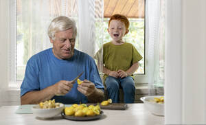Happy boy sitting near grandfather cutting fruits at home - MBLF00259
