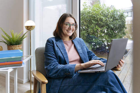 Smiling freelancer sitting with laptop on chair at home office - IHF01882