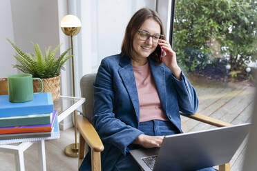 Lächelnde Geschäftsfrau, die mit einem Smartphone telefoniert und einen Laptop im Heimbüro benutzt - IHF01883