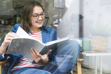 Happy freelancer examining reports sitting on chair at home office - IHF01884