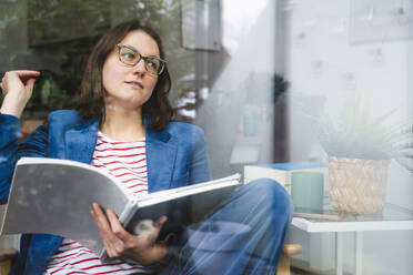 Thoughtful businessman holding book sitting at home office seen through glass - IHF01888