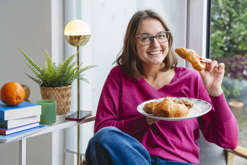 Smiling woman eating croissant sitting on chair at home - IHF01896