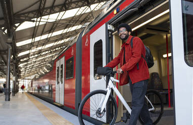 Lächelnder Mann, der mit seinem Fahrrad aus dem Zug am Bahnhof aussteigt - VRAF00376