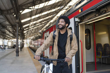 Lächelnder Pendler, der sein Fahrrad in der Nähe des Zuges am Bahnhof schiebt - VRAF00381