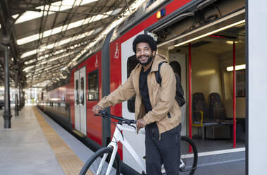 Lächelnder Pendler, der mit seinem Fahrrad aus dem Zug am Bahnhof aussteigt - VRAF00382