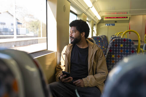 Mann mit Smartphone in der Hand und Blick aus dem Fenster in einem U-Bahn-Zug - VRAF00383