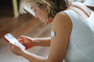 Blutzuckersensor am Arm einer Frau, die ihr Smartphone zu Hause benutzt - MDOF01809