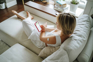 Woman with diabetes sensor using smart phone sitting on sofa at home - MDOF01812