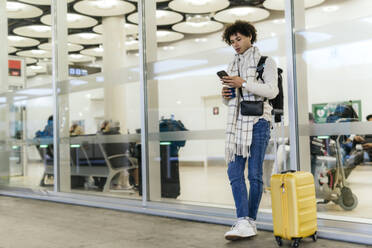 Junger Mann mit Koffer und Smartphone an einer Glaswand im Flughafen lehnend - PBTF00494