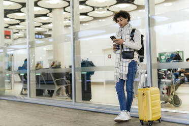 Young man with suitcase using smart phone leaning on glass wall at airport - PBTF00494
