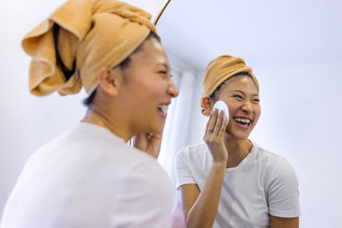 Fröhliche Frau reinigt ihr Gesicht mit einem Wattestäbchen vor dem Spiegel zu Hause - ASNF00020