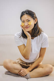 Lächelnde Frau mit Hautpflegeflecken unter den Augen, die zu Hause auf dem Sofa sitzt - ASNF00024