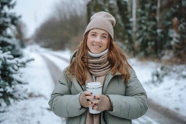 Lächelnde Frau mit warmer Kleidung und Thermoskanne in der Hand - NLAF00243