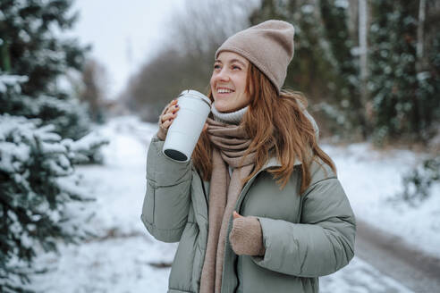 Glückliche Frau mit Thermoskanne auf einem Fußweg im Winter - NLAF00244