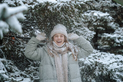 Glückliche Frau spielt mit Schnee unter einem Baum im Winter - NLAF00251