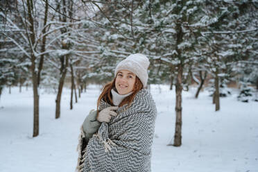 Smiling woman wrapped in blanket near trees on snow - NLAF00255