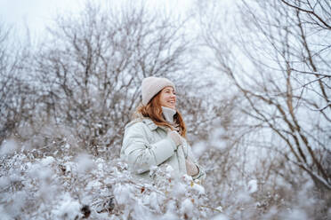 Glückliche Frau mit Strickmütze inmitten von Bäumen im Winter - NLAF00267