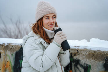 Smiling woman drinking tea from thermos near wall in winter - NLAF00268