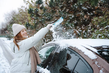Smiling woman cleaning snow from car roof in winter - NLAF00275