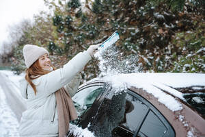 Lächelnde Frau reinigt im Winter den Schnee vom Autodach - NLAF00275