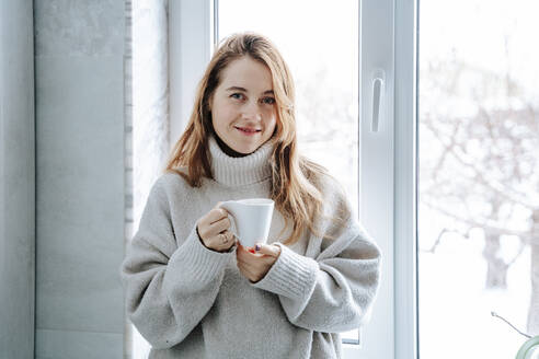 Lächelnde Frau mit braunem Haar hält Kaffeetasse in der Nähe von Fenster zu Hause - NLAF00278