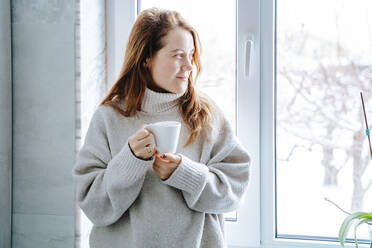 Lächelnde Frau hält Kaffeetasse in der Nähe von Fenster zu Hause - NLAF00279